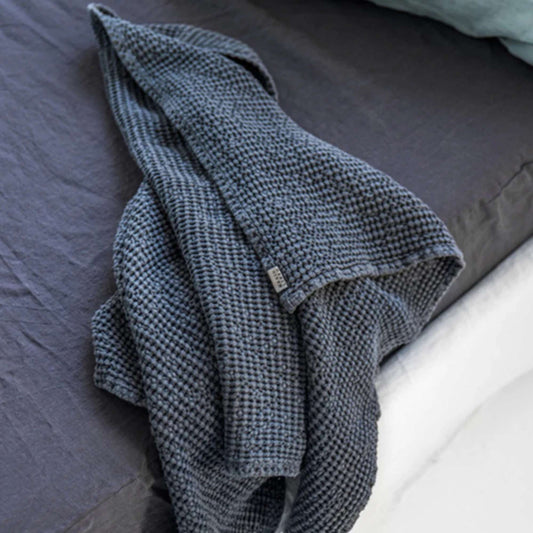 Mørkegrått håndkle i lin og bomull fra MagicLinen lagt på Woolroom sengetøy i lin og bomull, et rolig og naturlig uttrykk som fremhever teksturene og kvaliteten i materialene.