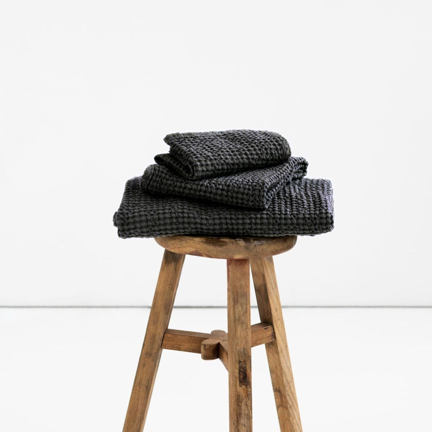 Mørkegrå håndklær i lin og bomull fra MagicLinen, et badehåndkle, et håndhåndkle og et ansiktshåndkle lagt på en krakk med naturlig lys som fremhever den myke teksturen og den rolige, luksuriøse følelsen.