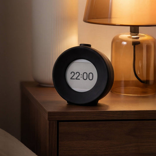 Digital vekkerklokke med E Ink skjerm på nattbord ved siden av lampe i rolig kveldsbelysning