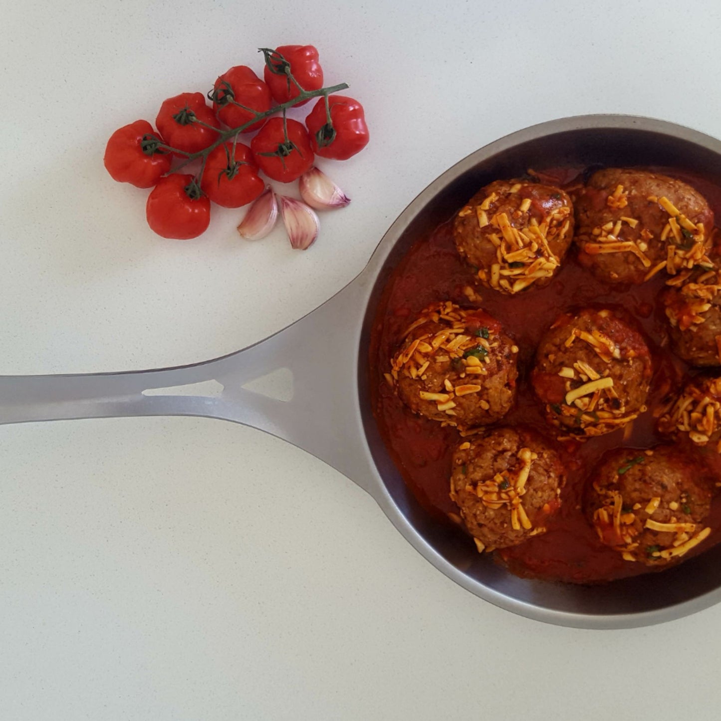 Kjøttboller i tomatsaus toppet med revet ost i en rustfri stålstekepanne, med friske tomater og hvitløk ved siden av.