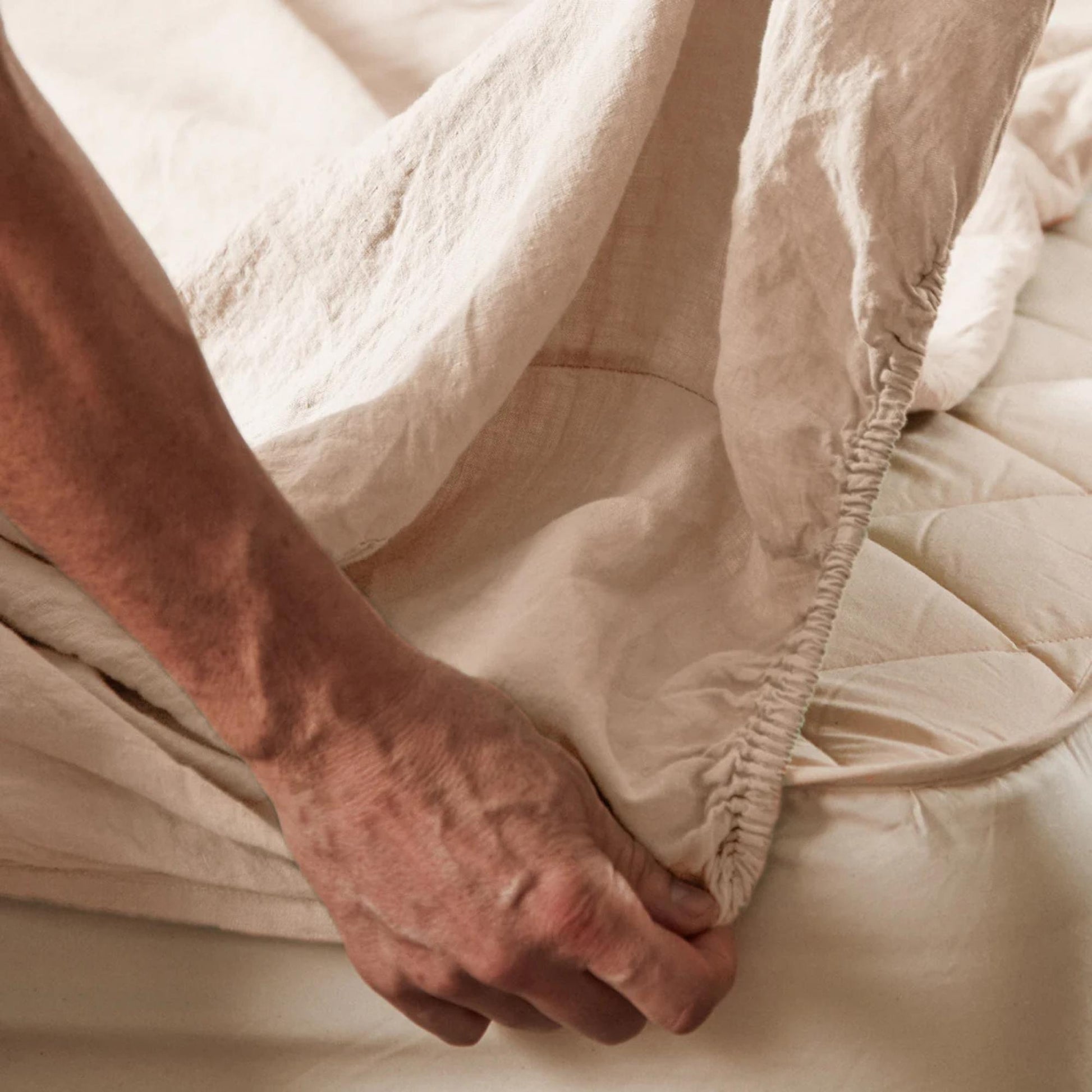 En mann legger på Mariana stretchlaken i økologisk bomull og lin i fargen sand, viser den elastiske kanten som gir perfekt passform