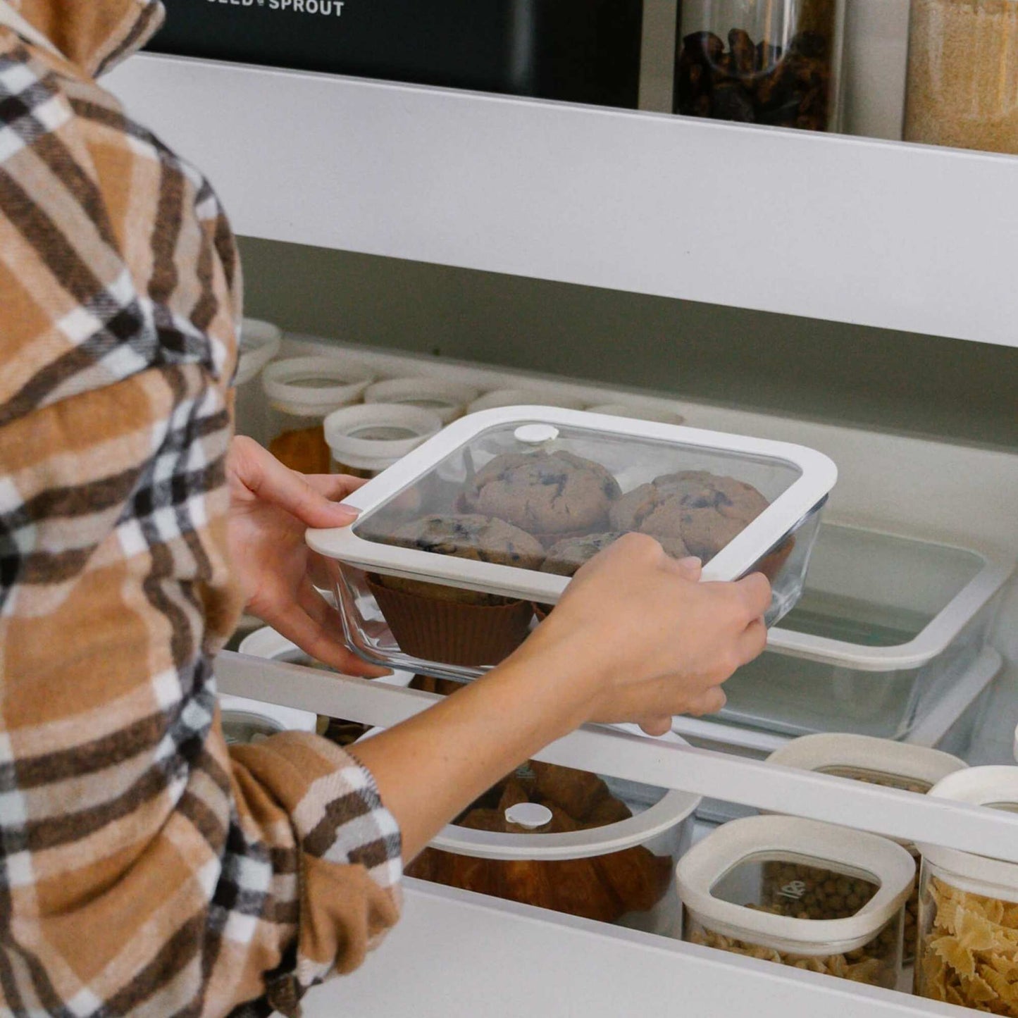 Person setter en glassbeholder med muffins inn i matskapet sammen med andre glassbokser.