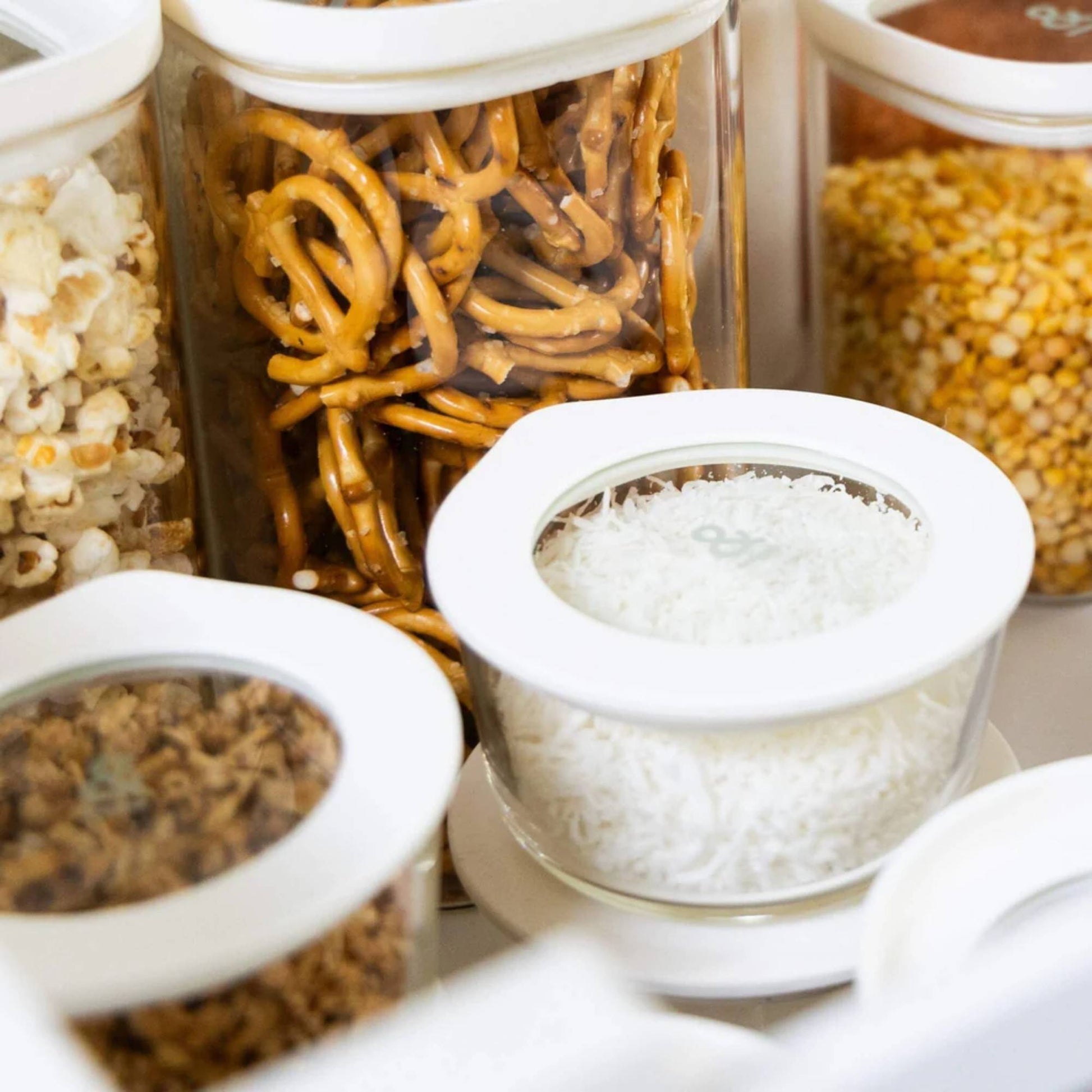 Glassbeholdere med glasslokk fylt med tørrvarer som ris, popcorn og pretzels, brukt til plastfri og oversiktlig kjøkkenoppbevaring.