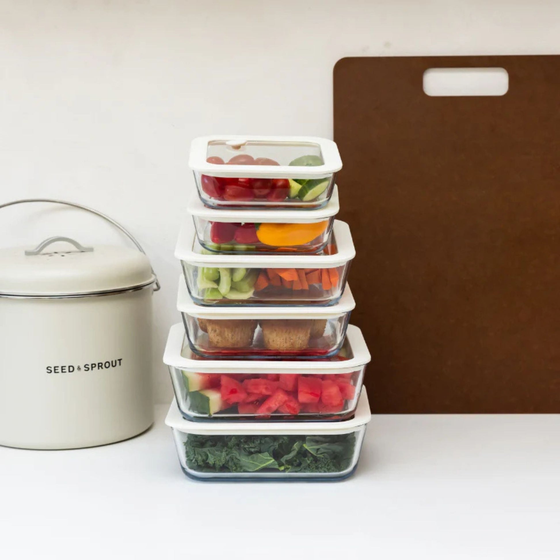 Stablede glassbeholdere med ferske grønnsaker og muffins, ved siden av en kjøkkenkompost og skjærebrett på benken.