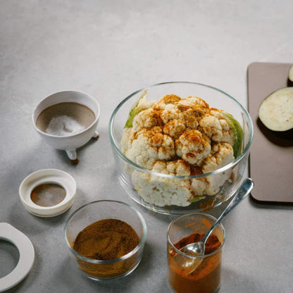 Glassbolle i borosilikatglass fylt med blomkål som krydres, omgitt av små skåler med krydder og ingredienser på kjøkkenbenk.