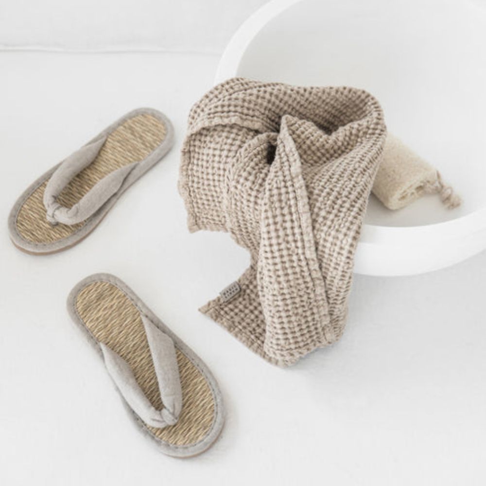 Badematte i lin og bomull i beige med vaffelstruktur, liggende på gulvet ved siden av to naturlige sandaler, i et lyst og rolig baderom