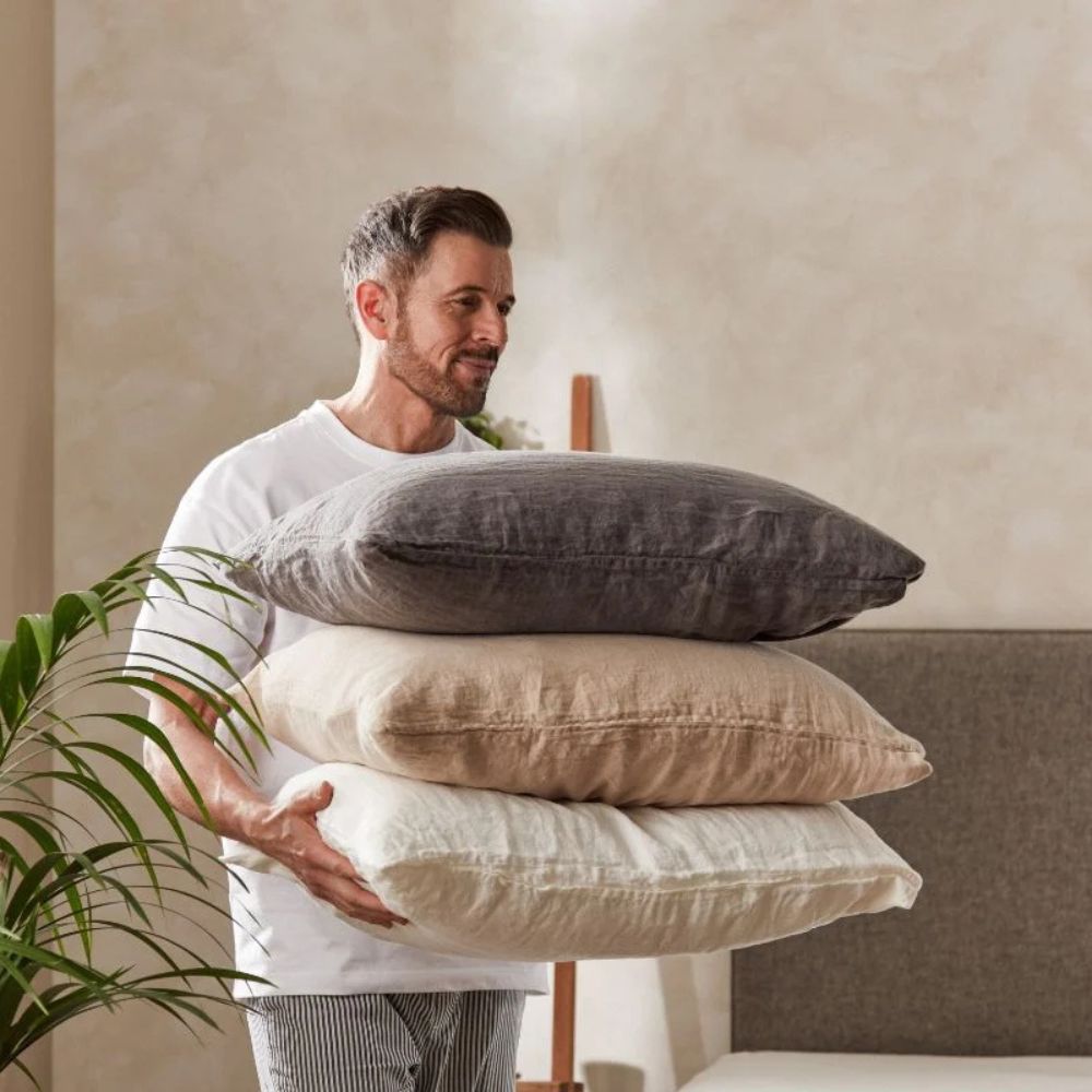 Mann bærer puter med Mariana sengetøy i naturlige farger av sand og mørkegrå, laget av lin og økologisk bomull