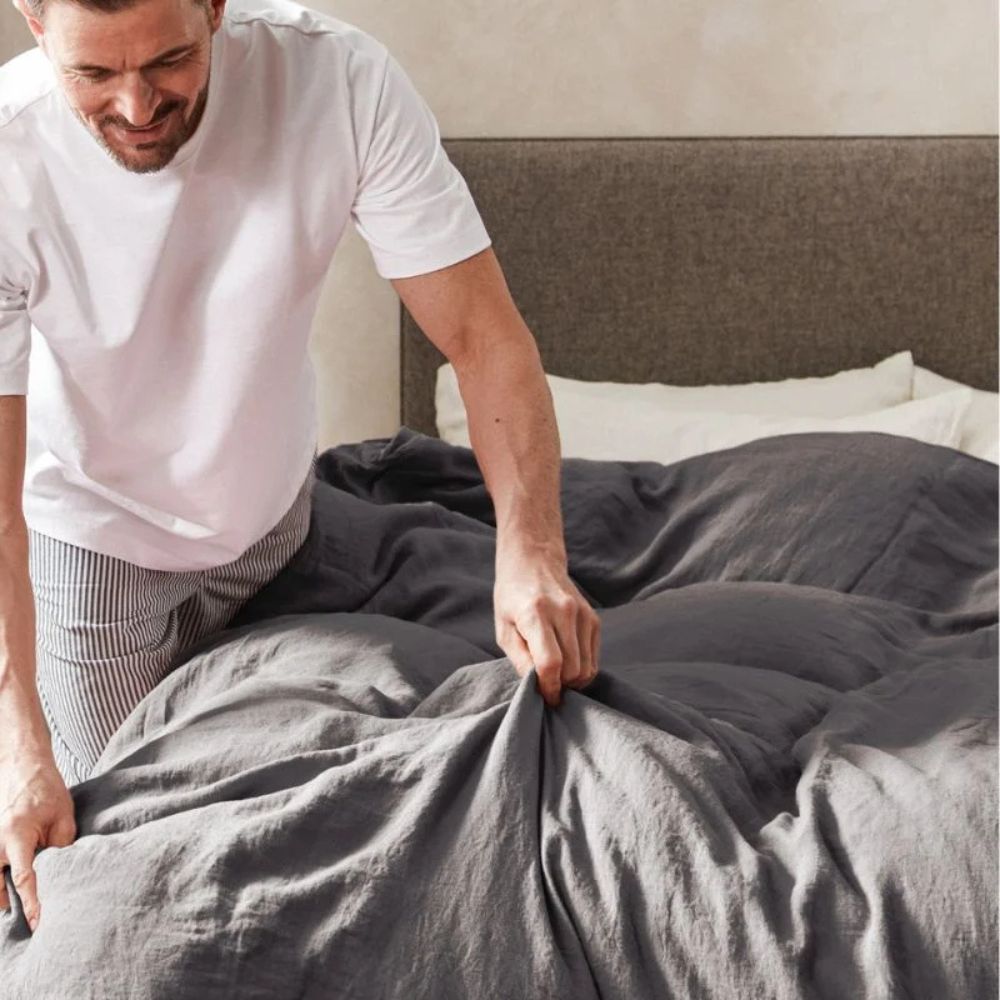 Mann rer opp sengen med Mariana sengesett i økologisk bomull og lin i fargen Charcoal – pustende og bærekraftig sengetøy for et sunnere hjem