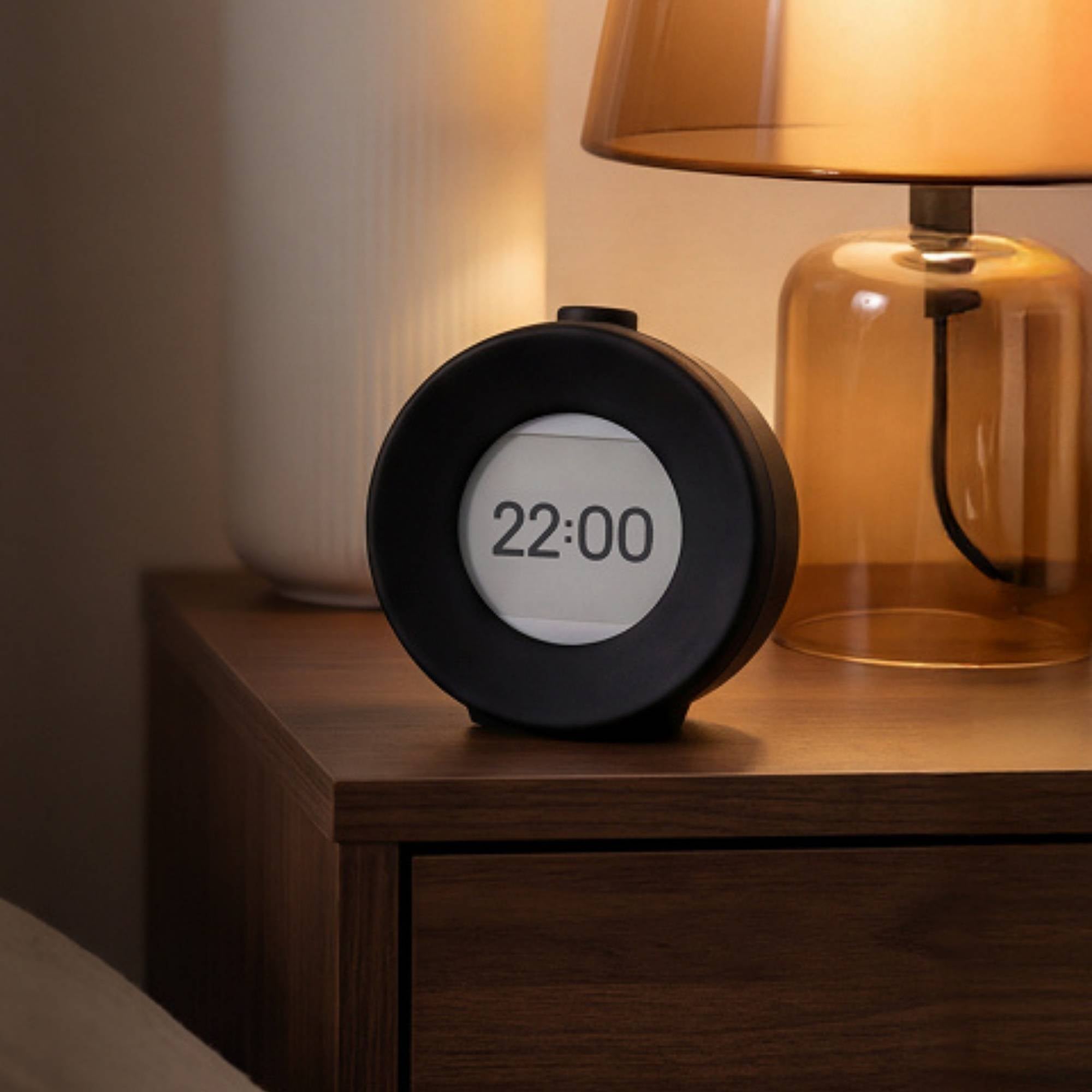 Digital vekkerklokke med E Ink skjerm på nattbord ved siden av lampe i rolig kveldsbelysning