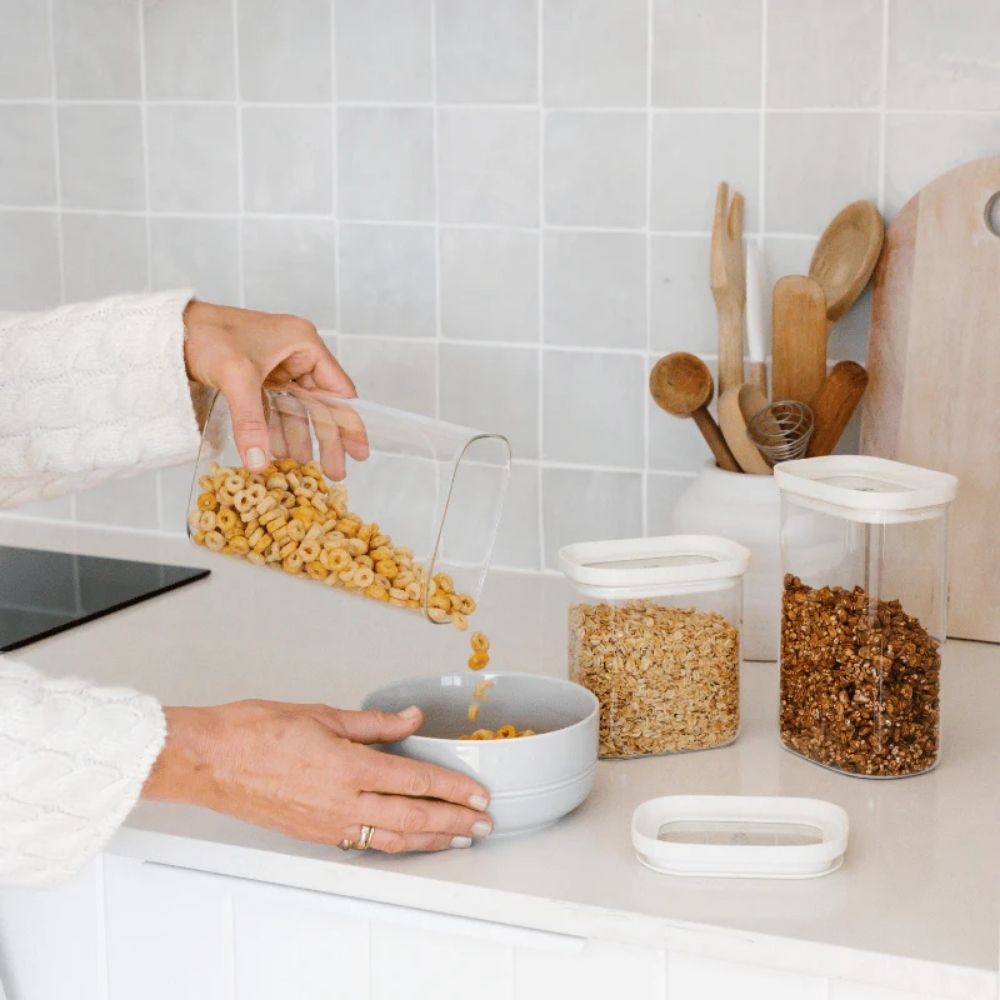 Person som heller frokostblanding fra en rektangulær glassbeholder med glasslokk i en bolle på kjøkkenbenken, omgitt av plastfrie oppbevaringskrukker fra Seed & Sprout.