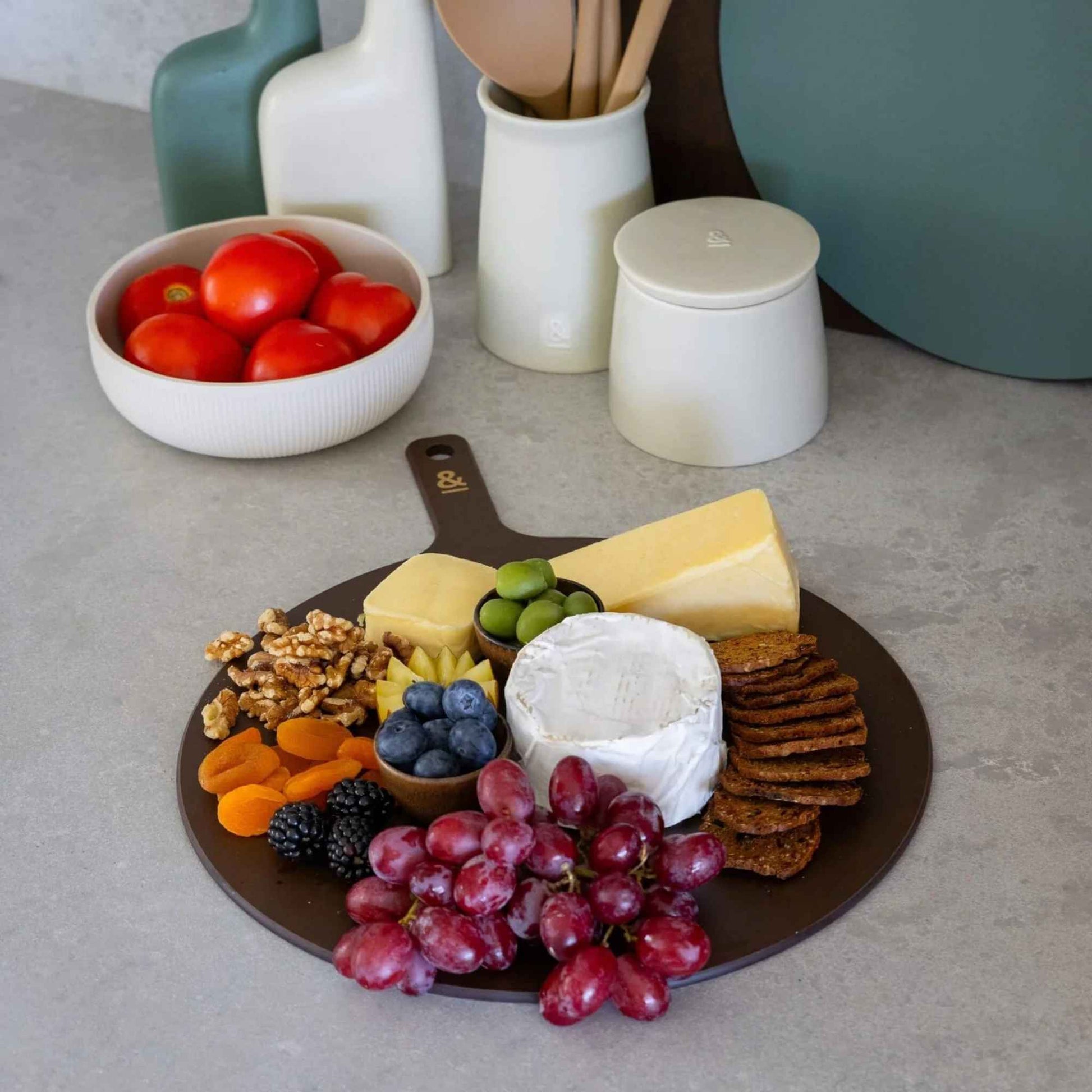 Rund skjærefjøl i trefiber i fargen Nutmeg brukt til servering av ost, kjeks, druer, nøtter og bær på kjøkkenbenk