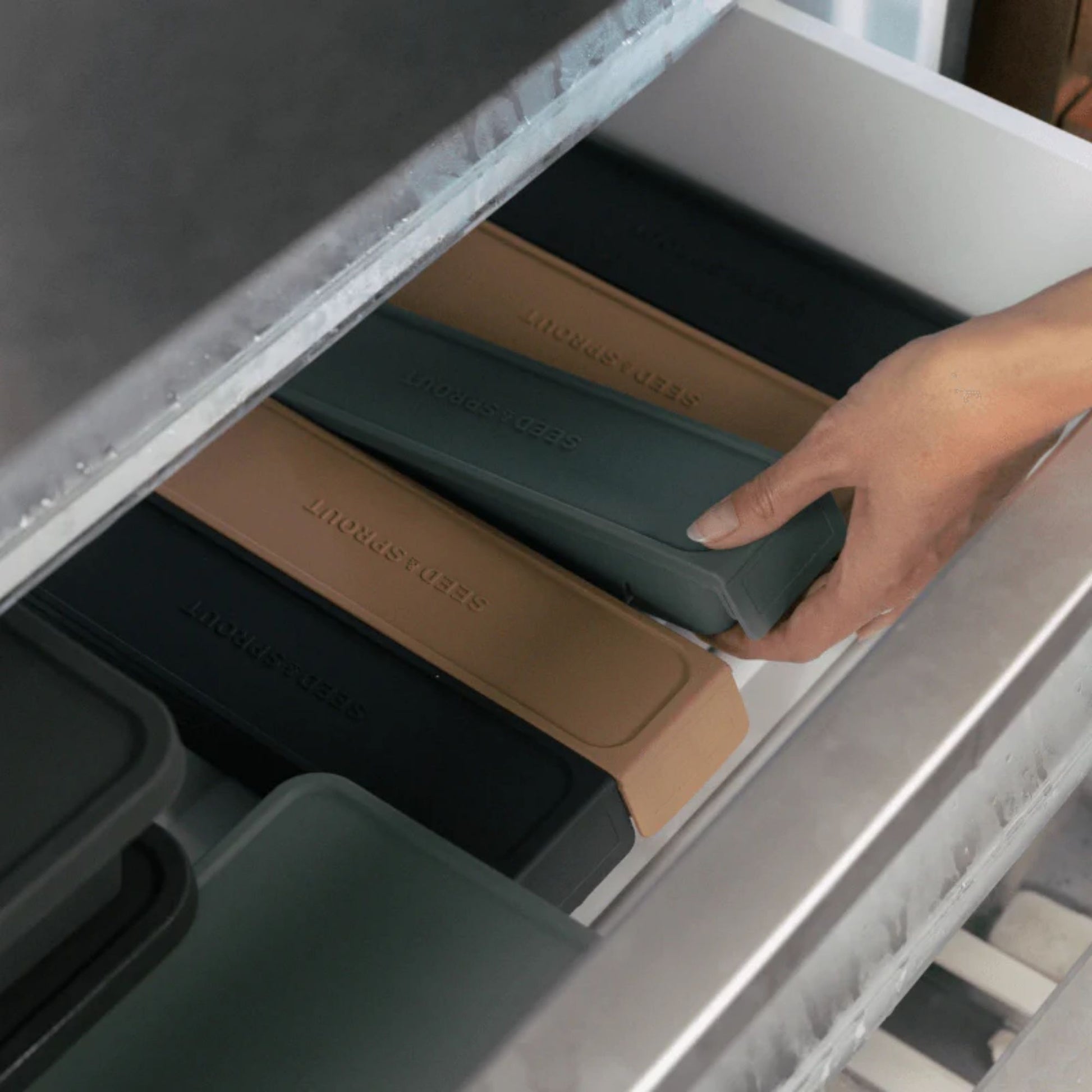 Stabelbare fryseformer i LFGB-sertifisert platinumsilikon fra Seed & Sprout organisert i en fryseskuff. Hånd som plasserer formen, viser hvor praktiske og plassbesparende de er.