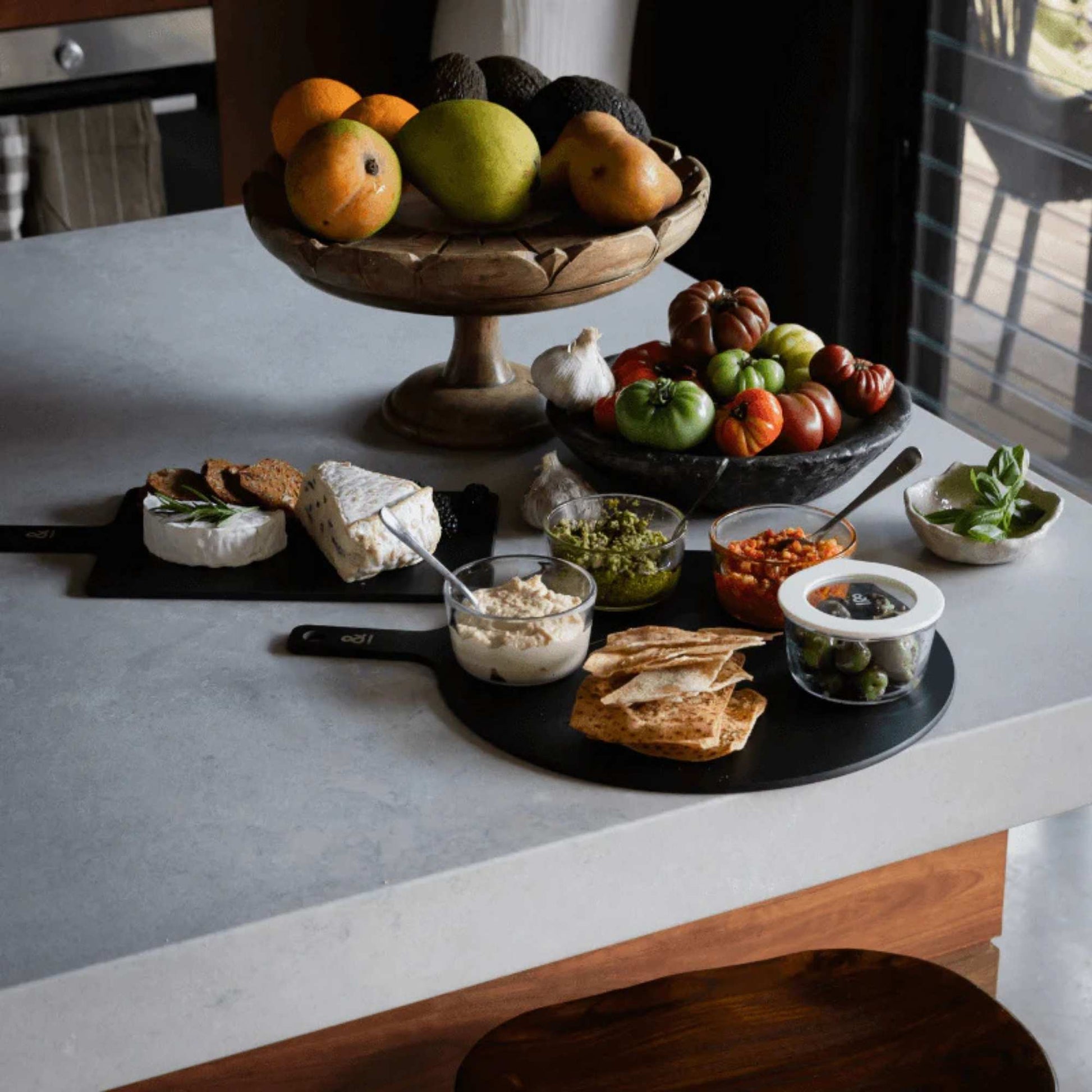 Glassboller med smørbare pålegg, dipper og kjeks pent arrangert på svart serveringsbrett på en lys kjøkkenbenk, med frukt og grønnsaker i bakgrunnen.