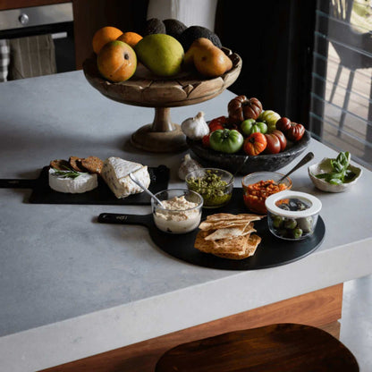 Glassboller med smørbare pålegg, dipper og kjeks pent arrangert på svart serveringsbrett på en lys kjøkkenbenk, med frukt og grønnsaker i bakgrunnen.