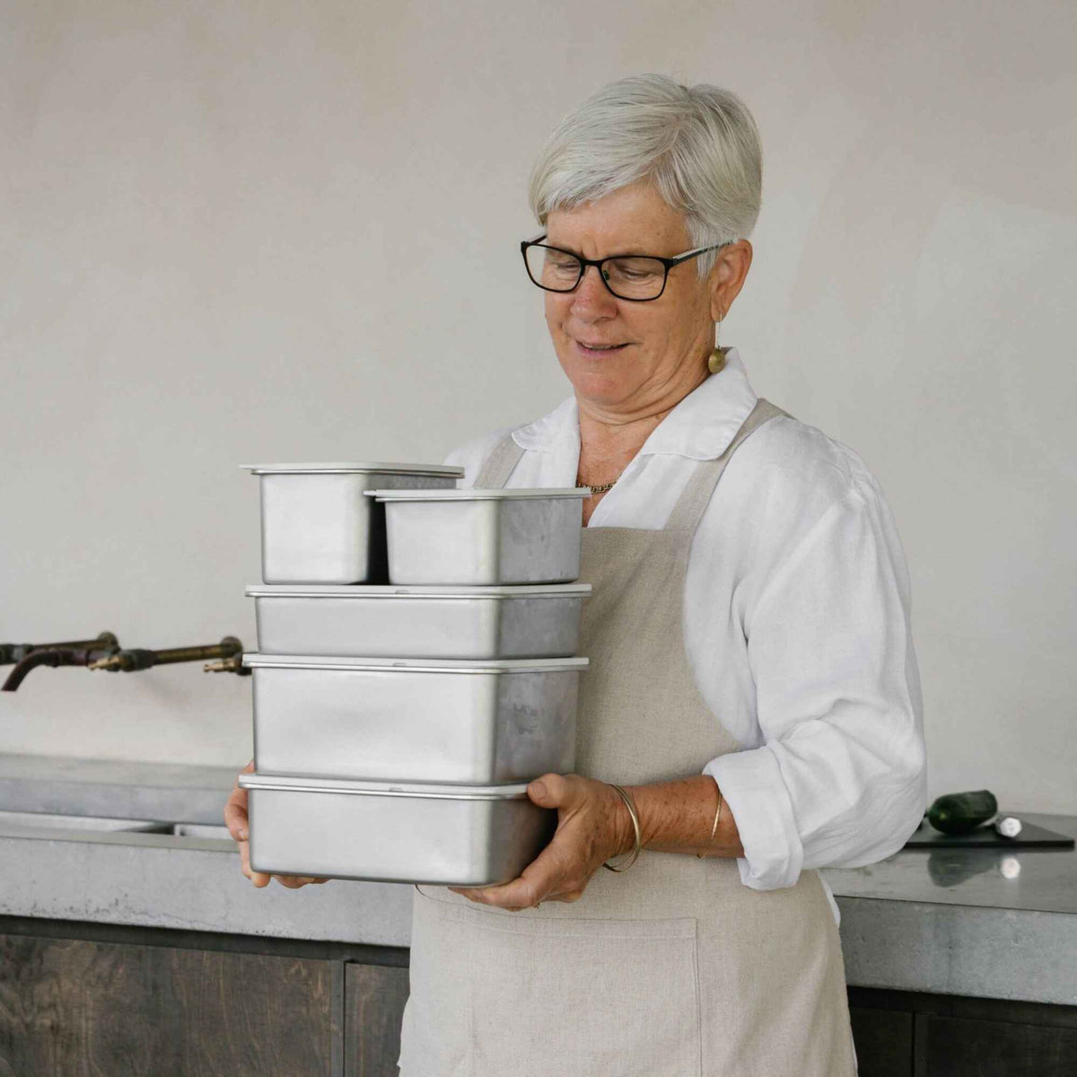 Person holder fem stablede, rektangulære matbeholdere i rustfritt stål med lokk fra Seed & Sprout på et kjøkken.