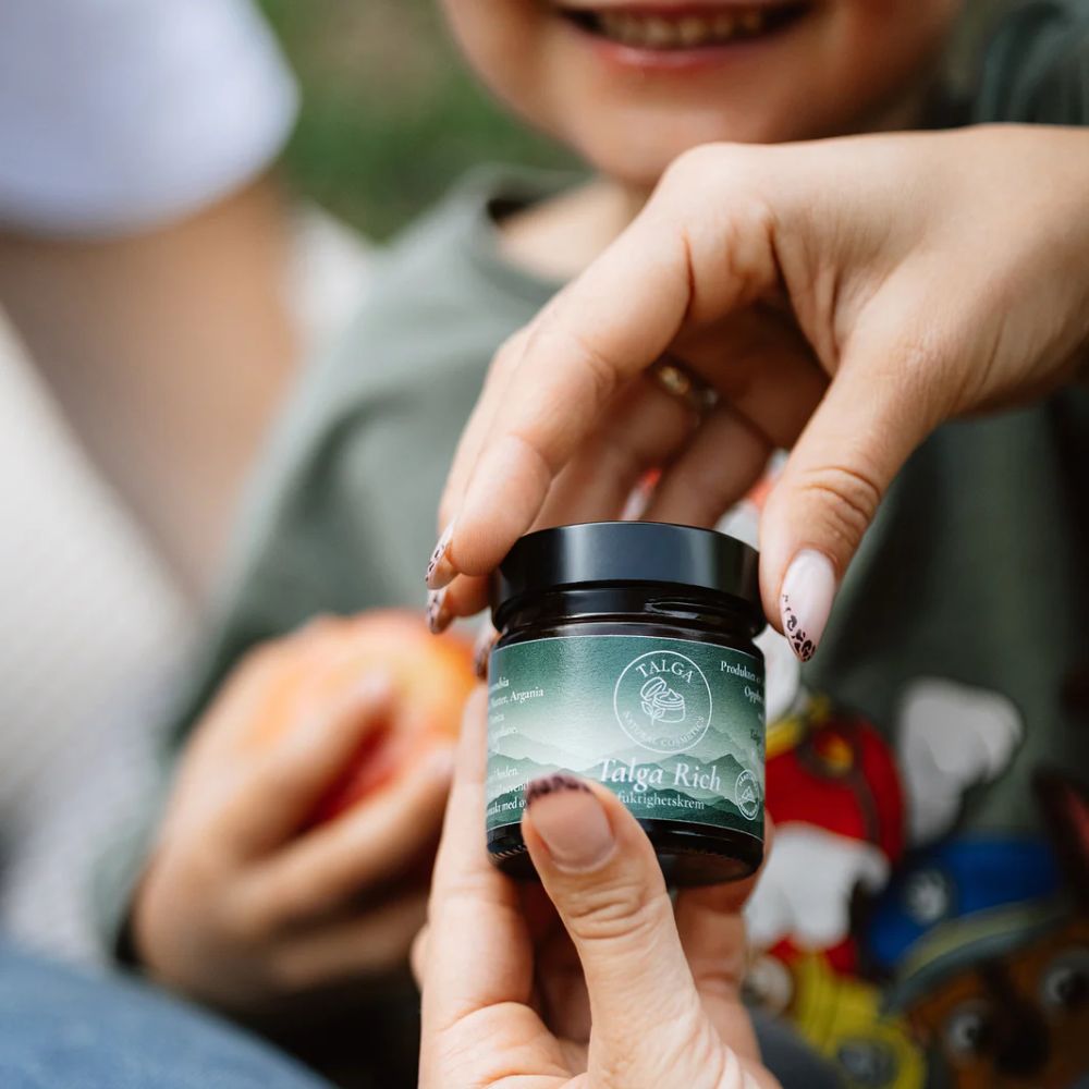 Voksen som åpner Talga Rich ansiktskrem mens et barn smiler i bakgrunnen – en mild, naturlig krem som også passer godt for familier og sensitiv hud.