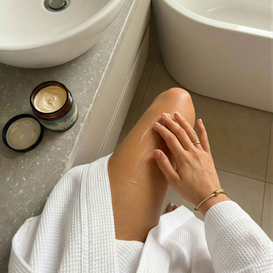 Person i hvit morgenkåpe smører Talga Rich Body Butter på benet på et lyst og rolig bad. Åpen krukke med kroppssmør står ved siden av vasken. Naturlig og nærende kroppspleie for myk og hydrert hud.