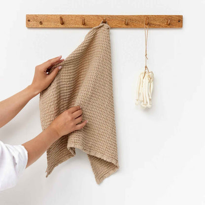 Beige vaffelvevd kjøkkenhåndkle i 53 % lin og 47 % bomull hengende på knaggrekke med en hånd som holder i håndkleet.