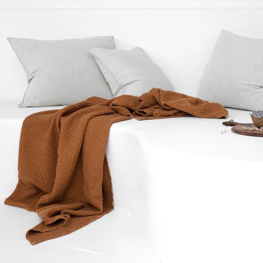 Waffle-pledd i lin og bomull i fargen cinnamon lagt på en utesofa, som viser den naturlige teksturen og den varme tonen i et rolig uteområde.