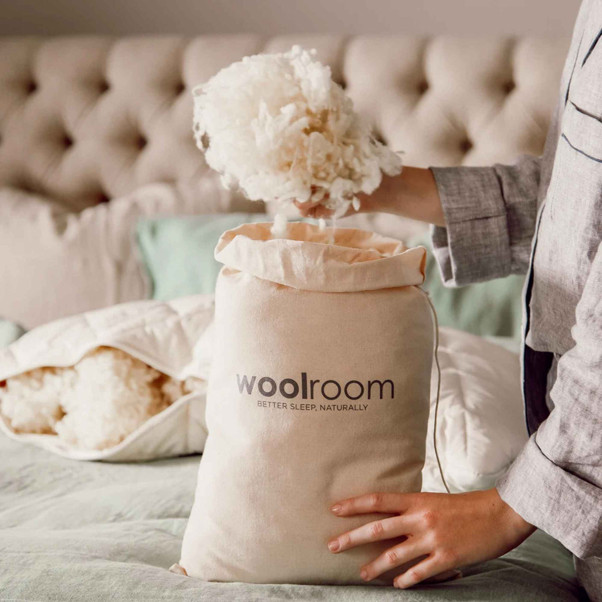 Person holder løst ullfyll over en Woolroom-stoffpose, med en åpen ullpute på sengen i bakgrunnen.